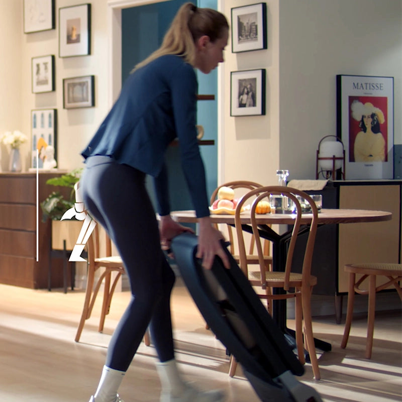Eine Frau in Sportkleidung stellt ein Kingsmith WalkingPad Z1 Klappbares Laufband für Schreibtisch in einem Essbereich mit Holzstühlen, gerahmter Wandkunst und einem Tisch mit Obst auf.