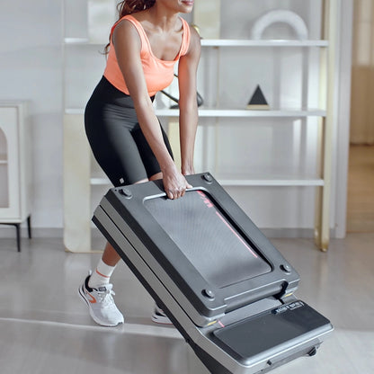 Eine Frau in Sportkleidung hebt ein Kingsmith WalkingPad Z3 Hybrid+ Klappbares Laufband für Schreibtisch in einem modernen, hellen Raum mit Regalen im Hintergrund.