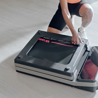 Eine Person kniet auf dem Boden und faltet ein schwarz-rotes Kingsmith WalkingPad Z3 Hybrid+ Klappbares Laufband für Schreibtisch zur platzsparenden Aufbewahrung in einem gut beleuchteten Raum.