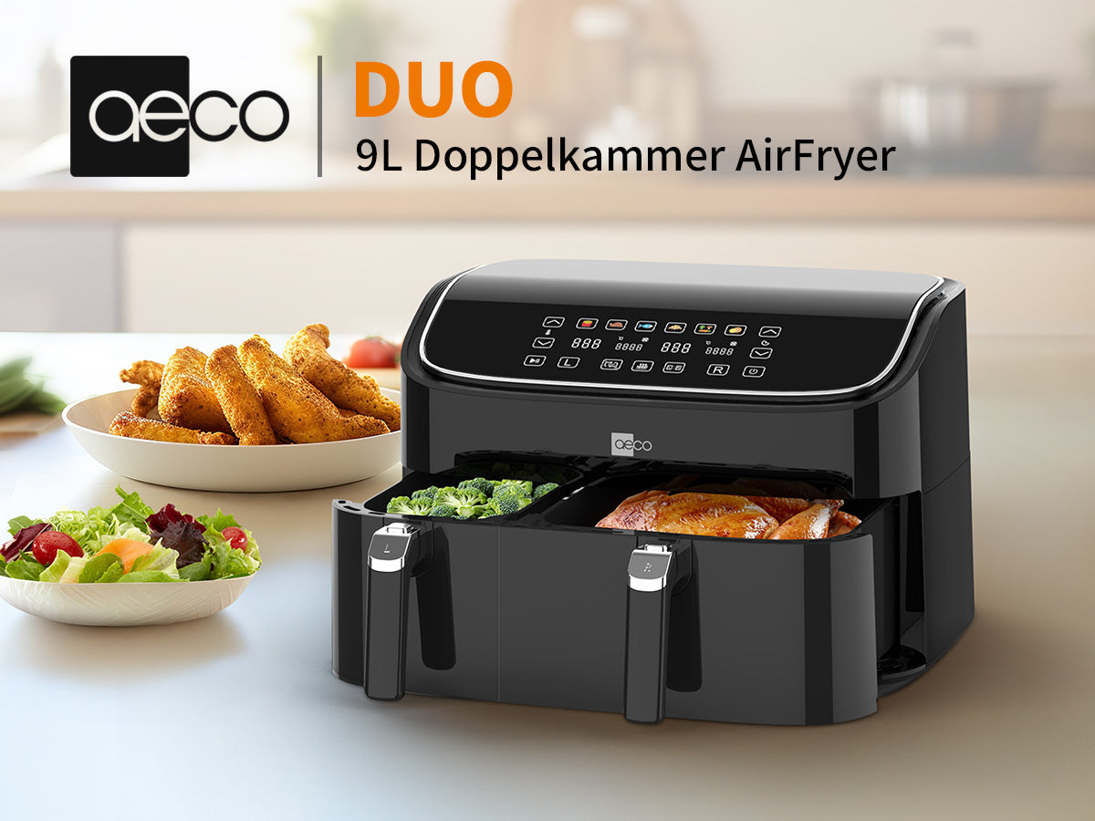 Eine schwarze aeco DUO Doppelkammer AirFryer mit 9 Liter Fassungsvermögen steht auf der Küchenarbeitsplatte, gefüllt mit gebratenem Hähnchen und Gemüse; im Hintergrund befindet sich ein Teller mit paniertem Essen.