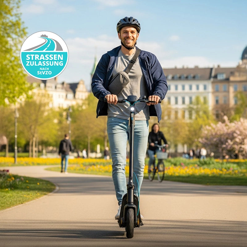 Ein Mann mit Helm fährt mit dem Odys ALPHA X5 PRO E-Scooter über einen Parkweg, der von blühenden Bäumen und städtischen Gebäuden gesäumt ist. Eine StVZO-Plakette mit Straßenzulassung und "Reichweite 50 km" sind die wichtigsten Merkmale.