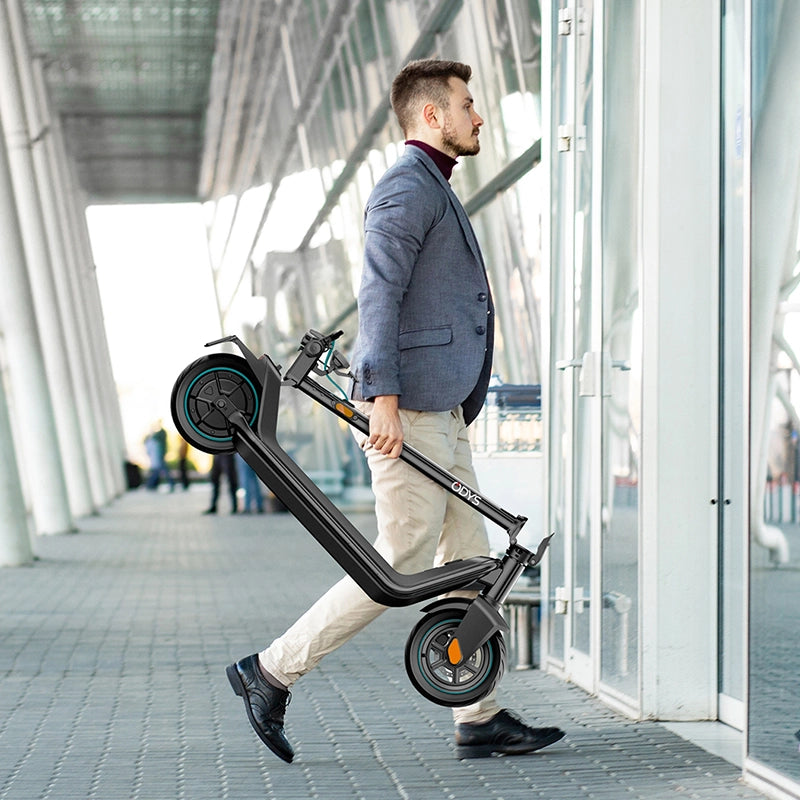 Ein Mann in einem Blazer trägt einen zusammengefalteten Odys ALPHA X5 PRO - E-Scooter, der für seine 50 km Reichweite bekannt ist, während er in ein modernes Glasgebäude geht.