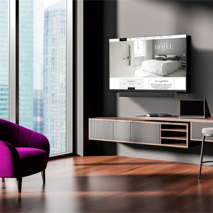 Ein modernes Wohnzimmer mit einem an der Wand montierten Dyon DYON Movie Smart 65 VX Smart TV (65", 164 cm), der eine Hotel-Website anzeigt, mit einem schwebenden Schrank, einem Laptop und einem magentafarbenen Sessel an den Fenstern mit Blick auf hohe Gebäude.