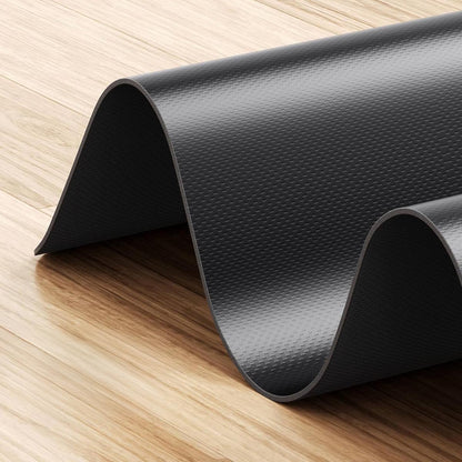 Eine rutschfeste Kingsmith WalkingPad-Laufband-Bodenmatte in strukturiertem Schwarz ist teilweise aufgerollt und liegt in sanften Wellen auf einem hellen Holzboden.