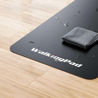 Eine rutschfeste Kingsmith WalkingPad-Laufband-Bodenmatte in Schwarz liegt auf einem Holzboden, auf der sich Wassertropfen und ein zusammengefaltetes graues Handtuch befinden.