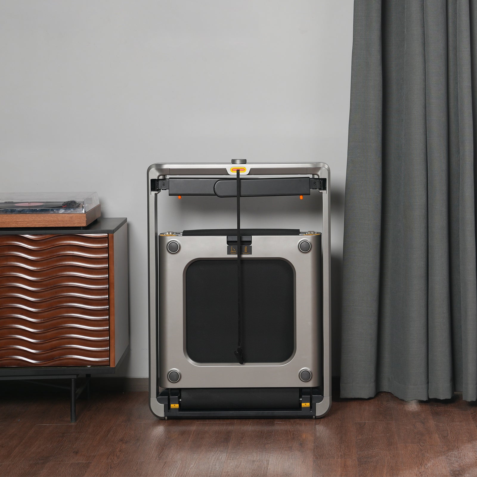 Das Kingsmith WalkingPad X214 Klappbares Laufband steht aufrecht an einer Wand neben einem modernen Holzschrank und einem grauen Vorhang auf einem Holzboden und unterstreicht das schlanke und platzsparende Design des Fitnessgeräts.