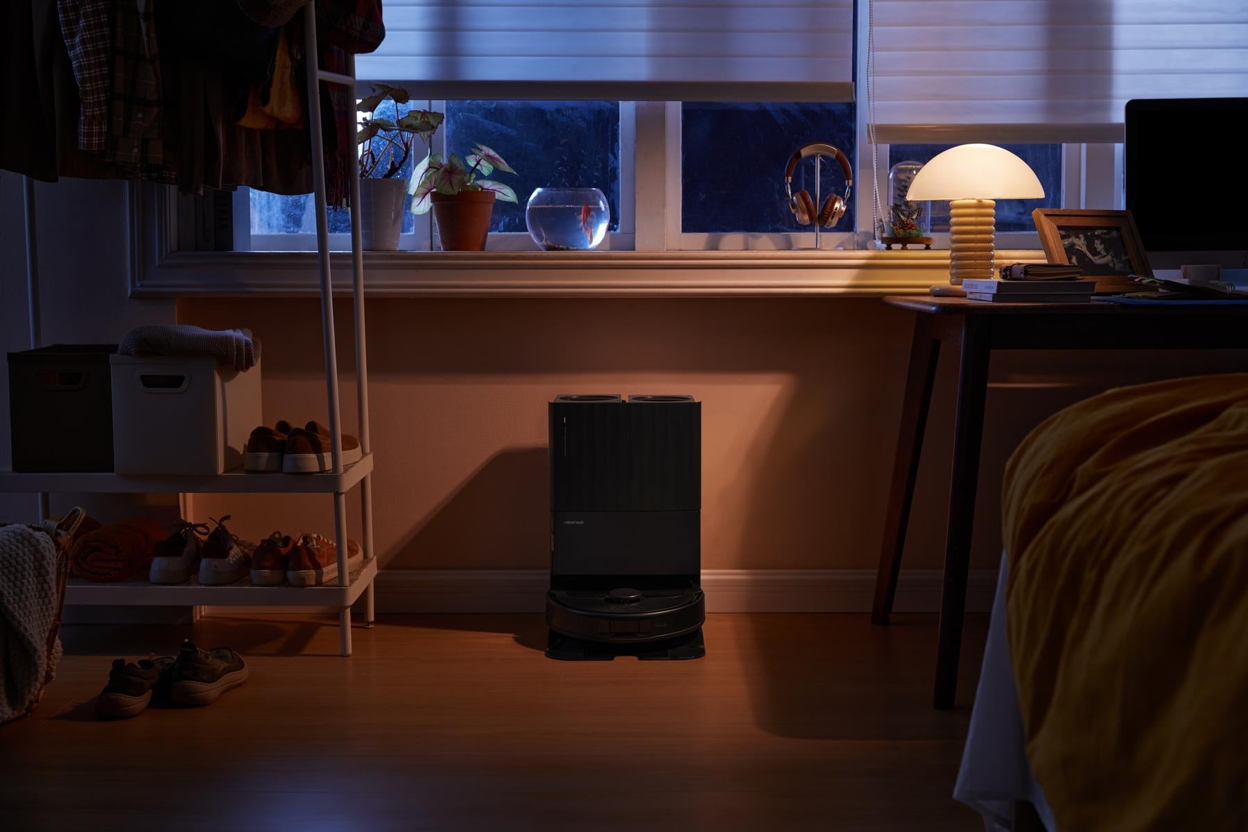 Ein Roborock Qrevo 598 Black ruht auf seiner multifunktionalen Dockingstation in einem schwach beleuchteten Schlafzimmer, mit einer leuchtenden Lampe, einem Bett, Regalen und einem Fenster im Hintergrund.
