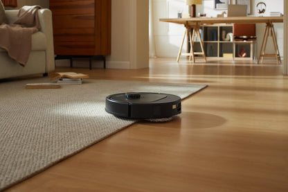 Ein schwarzer Roborock Qrevo 598 Staubsauger mit HyperForce-Saugfunktion wechselt in einem Wohnzimmer mit einem Sofa, einem Bücherregal und einem Schreibtisch im Hintergrund von Teppich auf Holzboden.