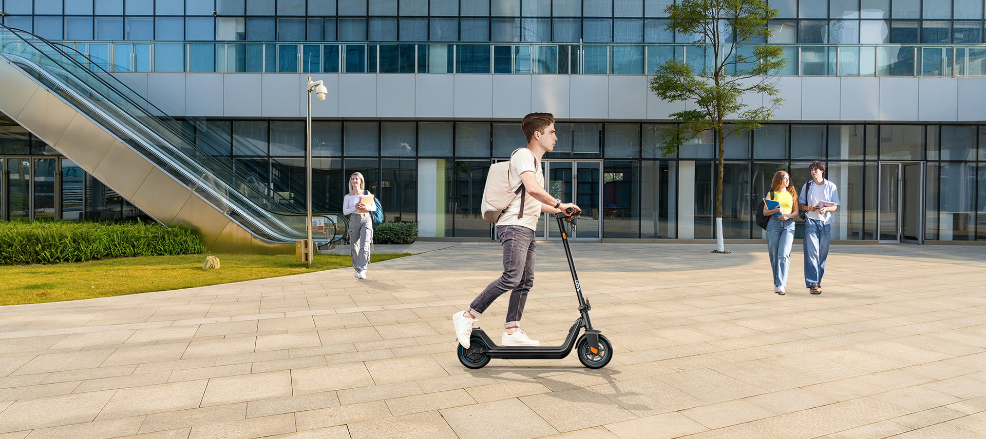 Ein junger Mann fährt mit einem Elektroroller auf einem gepflasterten Platz vor einem modernen Bürogebäude, während drei Personen in der Nähe spazieren gehen.