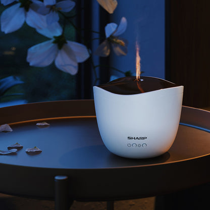 Der Sharp Ultraschall Aroma Zerstaeuber, ein weißer Luftbefeuchter und Aromadiffusor von Sharp, verströmt Nebel auf einem runden Tisch, mit weißen Blumen und einem Fenster im Hintergrund.