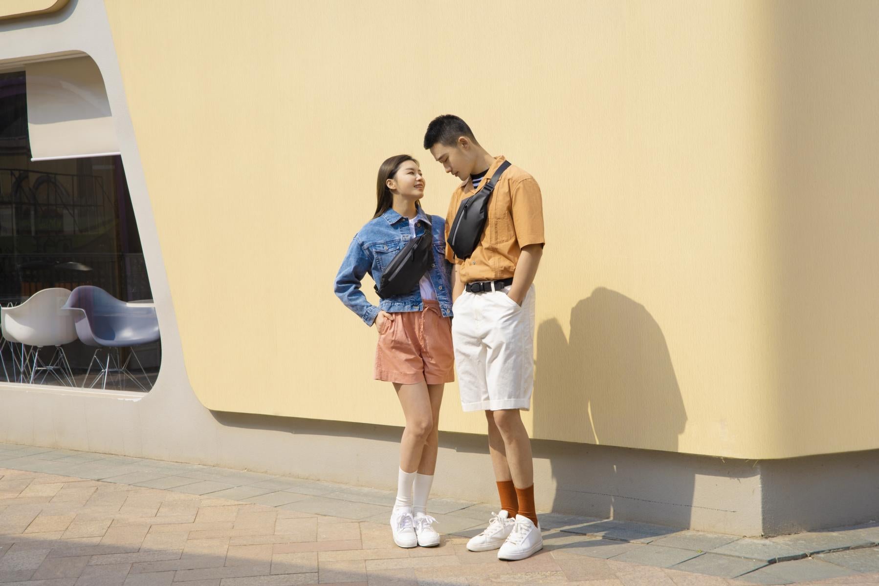 Ein junges Paar steht im Freien vor einer beigen Wand, trägt lässige Sommerkleidung mit Xiaomi Sports Fanny Packs, Turnschuhen und Shorts und sieht sich an.