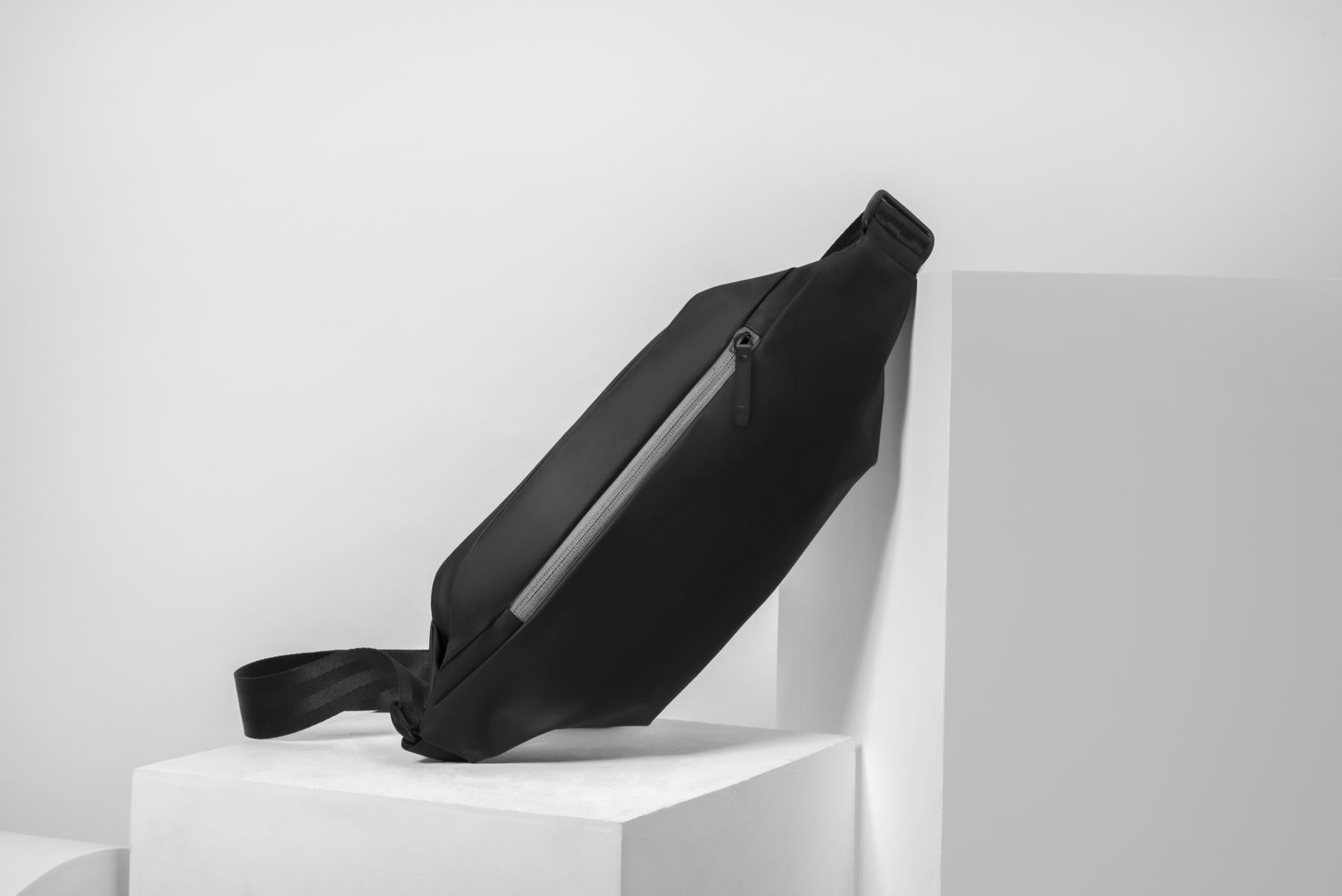 Die schwarze Xiaomi Sports Fanny Pack aus Kunstleder mit 2,25L Volumen und Frontreißverschluss steht vor einem weißen geometrischen Hintergrund.
